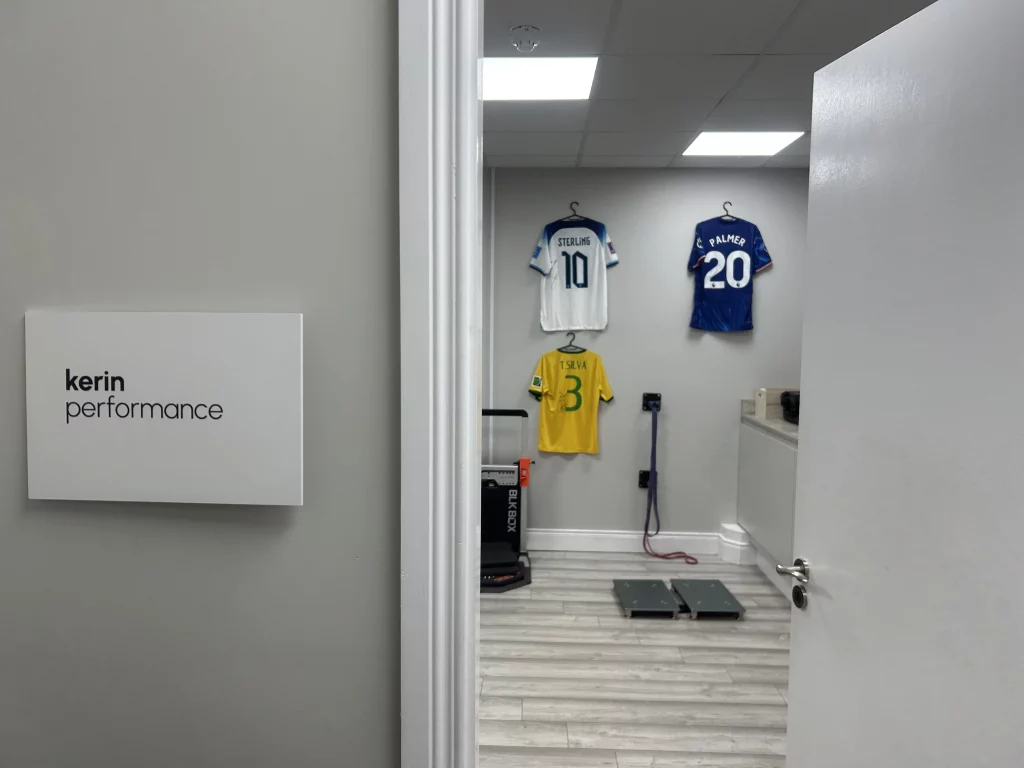 A room viewed through a doorway, with a sign reading "kerin performance" on the wall. Inside, three soccer jerseys with numbers 10, 3, and 20 hang on the back wall. The floor is wooden, and there are fitness equipment pieces visible.