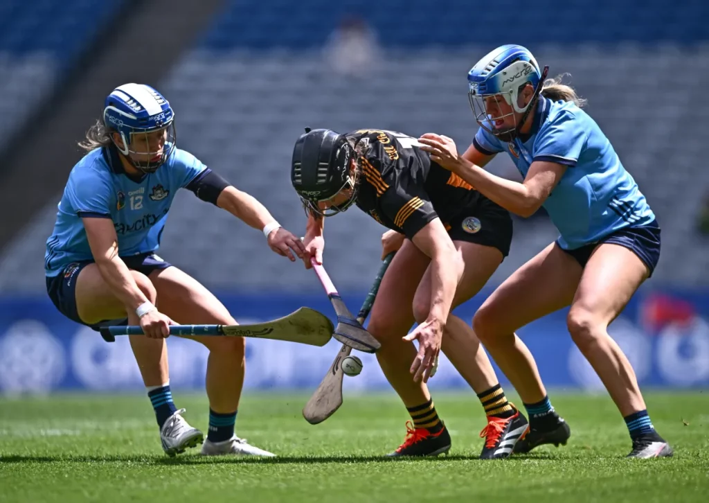 Three players in action on a field. One player in black and yellow is bent forward, attempting to control a ball, while two players in blue try to block her. All are wearing helmets and holding hurleys. The background shows an empty stadium.