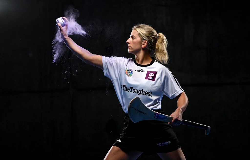 A woman in a sports uniform prepares to hit a ball with a hurley stick. She stands with her arm extended, holding the ball, as white dust billows around it. The background is dark, highlighting her focused expression and athletic stance.