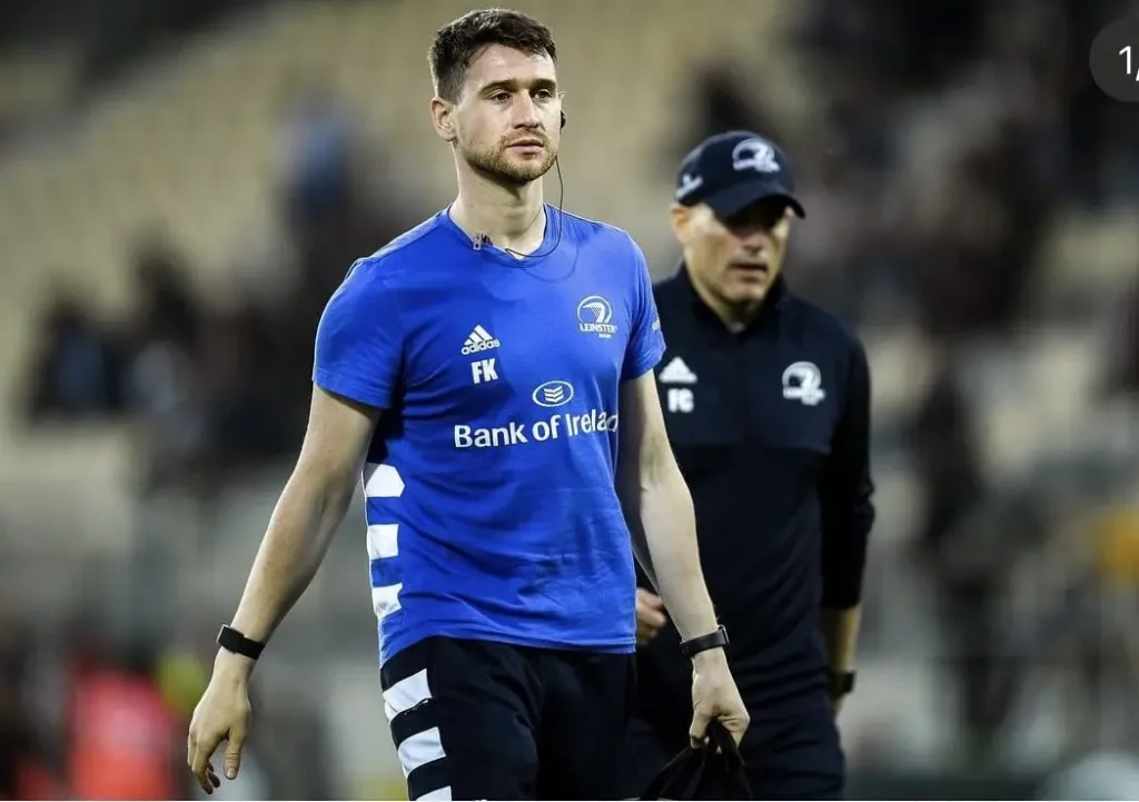 Fearghal in a blue sports shirt walks on a field, holding a bag. Another person in a sports jacket and cap walks nearby. The background shows a stadium with blurred seats.