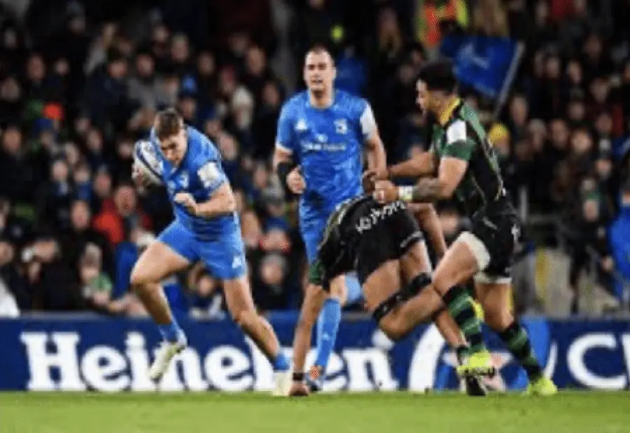 A rugby player in a blue jersey runs with the ball, evading two opponents in green and black. Two teammates in blue follow closely behind. The action takes place on a sports field with spectators in the background.