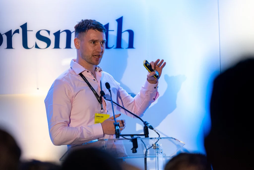 A man in a pink shirt stands at a transparent podium, giving a presentation. He holds a remote in one hand and gestures while speaking. A microphone and a blurred logo are visible in the background. The setting appears to be a conference or seminar.